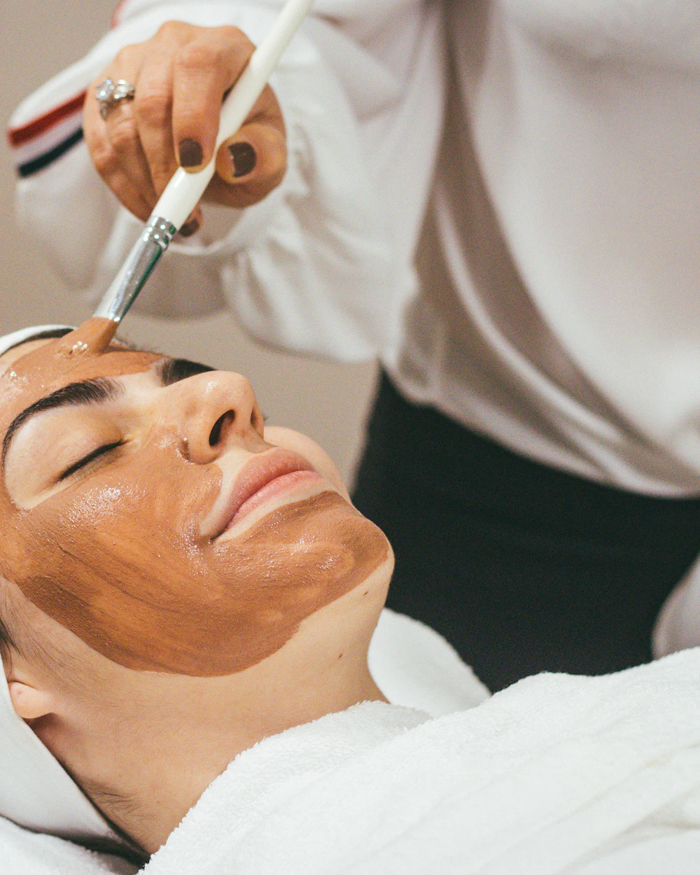 Client relaxing during a facial treatment while a clay mask is brushed onto her skin