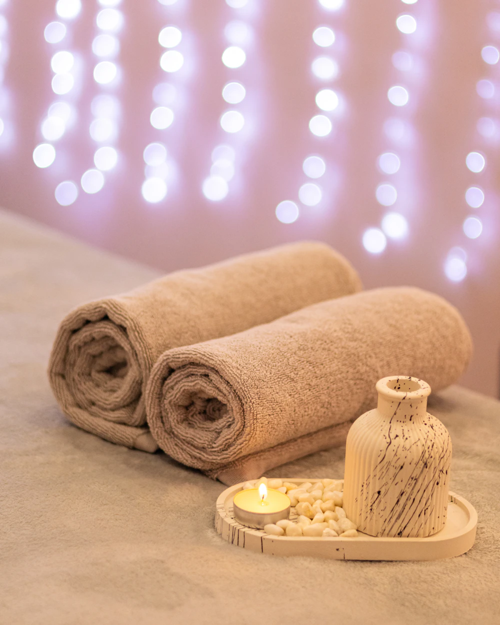 Rolled spa towels, a lit candle and warm string lights set up for a beauty treatment